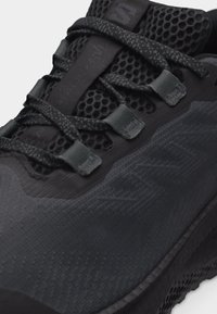 Κοντινή λήψη ενός μαύρου αθλητικού παπουτσιού με υφασμάτινο διχτυωτό επάνω μέρος, μαύρα κορδόνια και υφασμάτινα περάσματα κορδονιών με διακριτικές ανάγλυφες λεπτομέρειες επωνυμίας.