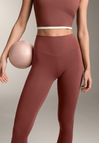 Primo piano di una persona che indossa leggings a vita alta color rosa e un top corto, mentre tiene una palla da ginnastica rosa tra il braccio e la vita.