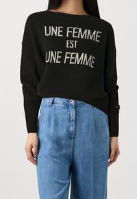 Svart stickad tröja med texten "UNE FEMME EST UNE FEMME" i ljusgrå. Har en avslappnad passform och ribbade muddar. Matchad med ljusblå denimbyxor.