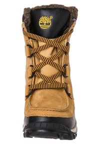 Hnědá kožená bota s černou gumovou podrážkou, vybavená chlupatou podšívkou, žlutým logem na patě a kříženými černými a žlutými tkaničkami.