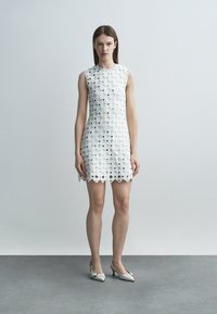 Vrouw die recht vooruit staat, gekleed in een mouwloze witte kanten jurk met geschulpte randen en witte hakken, tegen een effen lichtgrijze achtergrond.