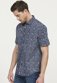Chemise en coton à manches courtes, présentant un fond bleu marine avec de petits motifs floraux blancs et un col boutonné.