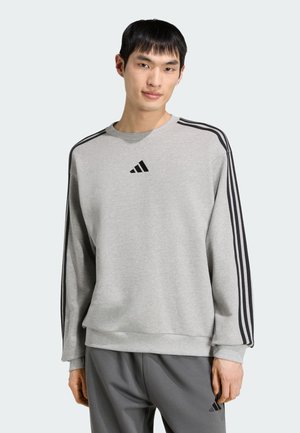 Mann trägt ein graues Adidas Sweatshirt mit schwarzen Streifen an den Ärmeln und passende graue Adidas-Hose, stehend vor einem hellen Hintergrund.