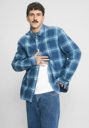 Mężczyzna z wąsami i tatuażami, ubrany w niebieską koszulę w kratę na guziki, nałożoną na białą koszulkę, oraz niebieskie dżinsy, pozujący na jednolitym tle.