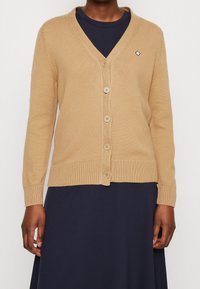 Beige cardigan med V-hals og ribbede ærmeafslutninger. Har fem knapper ned foran og et lille logodetalje på venstre bryst.