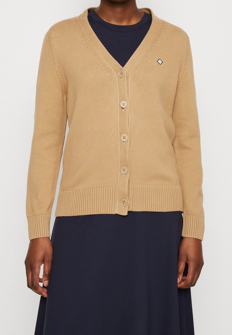 Beige cardigan med V-hals og ribbede ærmeafslutninger. Har fem knapper ned foran og et lille logodetalje på venstre bryst.