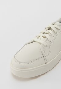Sneaker in pelle bianca con punta arrotondata, superficie texturizzata e lacci piatti. Dettagli cuciti visibili sulla parte superiore e sui lati.