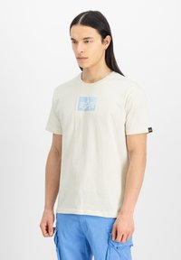 Cremefarvet bomulds T-shirt med et lyseblåt grafisk logo i midten. Korte ærmer, afslappet pasform og et lille mærke på venstre ærme.