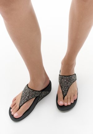 Pieds portant des tongs noires avec des sangles scintillantes sur un fond blanc, montrant une vue latérale et avant des sandales.