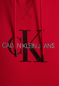 Sweat à capuche rouge avec grand logo noir "CK" et texte argenté "CALVIN KLEIN JEANS" sur la poitrine, avec cordons rouges à embouts noirs.