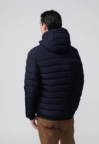 Uomo che indossa un giubbotto trapuntato nero con cappuccio e pantaloni marroni, in piedi con le spalle verso la telecamera su uno sfondo grigio uniforme.