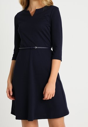 Vrouw draagt een donkerblauwe jurk tot op de knie met driekwart mouwen en een smalle bijpassende riem in de taille.