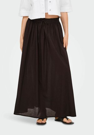 Woman wearing a long black drawstring skirt paired with black sandals and a cropped white buttoned shirt, standing against a plain background.