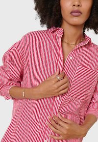 Red and white striped shirt featuring a button-down collar, long sleeves, and a front pocket with horizontal stripes. Cotton material.