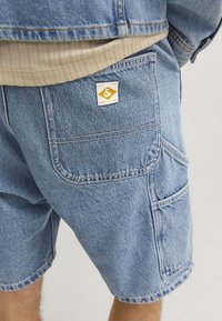 Denim shorts in light blue with visible stitching, multiple pockets, and a small yellow and white logo tag on the back pocket.