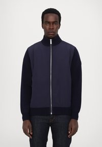 Navy bomber jacket with a high collar, zippered front, textured knit sleeves, and ribbed hem, paired with dark jeans.