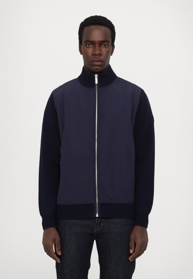 Navy bomber jacket with a high collar, zippered front, textured knit sleeves, and ribbed hem, paired with dark jeans.