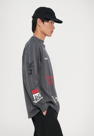 Jovem com boné preto, camisa de manga longa cinza com gráfico e texto vermelho e branco, calças pretas, de lado com as mãos nos bolsos.