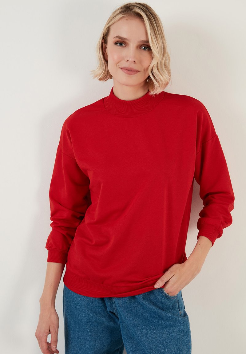 Sudadera roja con cuello alto y puños acanalados. La tela parece suave y tiene un corte relajado. Combinada con pantalones de denim azul claro.