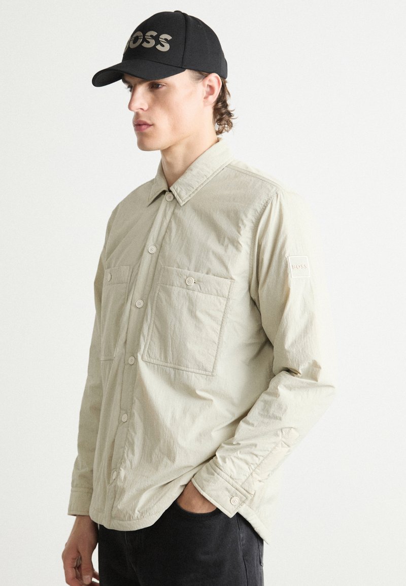 Beige lightweight shirt with two front pockets, button closure, and a soft texture, paired with a black cap featuring "BOSS" in silver lettering.