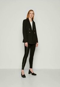 Μαύρο σακάκι διπλής σειράς με χρυσά κουμπιά, φορεμένο πάνω από λευκό τοπ, σε συνδυασμό με γυαλιστερά μαύρα κολάν και μαύρα παπούτσια με χοντρό τακούνι.