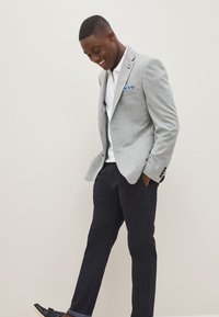 Blazer gris clair avec un mouchoir de poche bleu, polo blanc, pantalon foncé et mocassins noirs. Coupe ajustée avec une texture subtile.