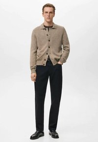 Beige gebreid vest met een kraag, knopen aan de voorkant, geribbelde manchetten, gecombineerd met zwarte broek en zwarte schoenen. Eenvoudig ontwerp.