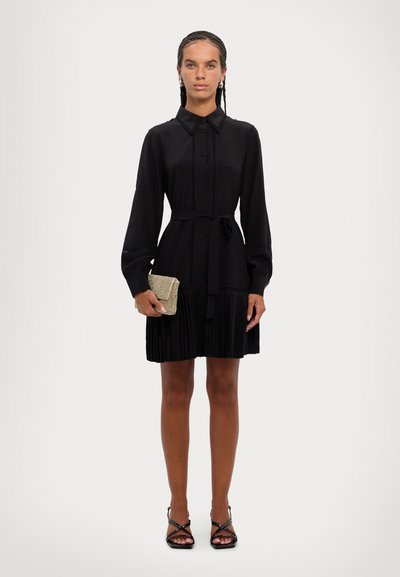Vestido negro con cuello y botón en la parte delantera, con mangas largas y falda plisada. Un par de tacones negros con tiras y un bolso metálico.