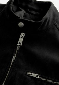 Chaqueta de ante negra con un cuello alto, que presenta un destacado zipper plateado y un bolsillo lateral con cremallera con un acento metálico.