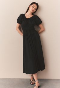 Schwarzes Maxikleid mit Puffärmeln und gerafftem Oberteil. Glatter Stoff fällt bis zum Knöchel. Modell steht mit Händen in der Hüfte und trägt Riemchensandalen.