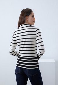 Chemise à manches longues rayée noire et blanche avec un col rond, confectionnée en tissu lisse, design ajusté, portée avec un jean bleu foncé.