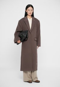 Smeđi oversized kaput od vune s ovratnikom, bočnim džepom i čistom siluetom. Model drži crnu torbu, nosi svijetlu majicu i bež hlače.