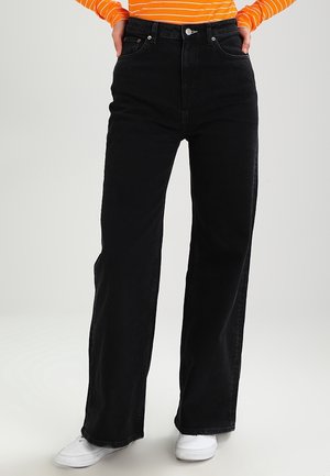 Person, die schwarze, hoch taillierte, weit geschnittene Jeans, ein orange-weiß gestreiftes Langarmshirt und weiße Turnschuhe trägt, die Hände in den Taschen.