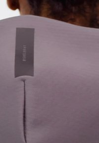 Top sportivo viola chiaro con una cucitura centrale e un'etichetta grigia con il testo "SPOTSHER", realizzato in un tessuto elastico e liscio.