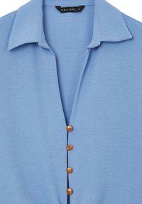 Camicetta azzurro chiaro con colletto e cinque bottoni marroni sul davanti. Il tessuto ha una consistenza liscia e morbida.