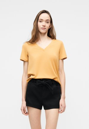 Kvinne med langt brunt hår som har på seg en sennepsgul T-skjorte med V-hals og svarte shorts med knyting i livet, stående foran en hvit bakgrunn.