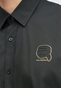 Crna košulja na kopčanje od glatkog tkanja s crno-zlatnim crtanim logotipom na prsima. Dugmad su crna s mat završetkom.