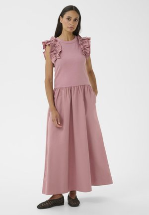 Femme portant une robe longue rose sans manches avec des détails à volants aux épaules et des chaussures noires tissées sans lacets, debout avec une main dans la poche.