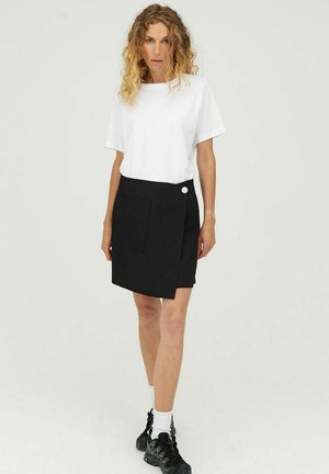 Jupe portefeuille noire avec une finition texturée, dotée d'une poche avant et d'un détail de bouton blanc, accompagnée d'un t-shirt blanc à manches courtes.