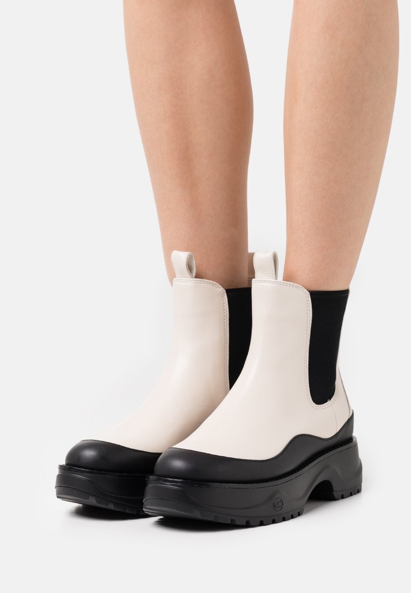 Bottines plateformes épaisses créme et noires avec languettes, portées sur des jambes nues devant un fond blanc.