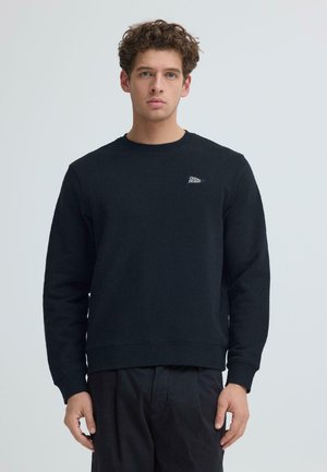 Giovane con capelli ricci indossa una felpa nera con piccolo logo bianco "Casual Friday" sul petto, in piedi davanti a uno sfondo chiaro uniforme.