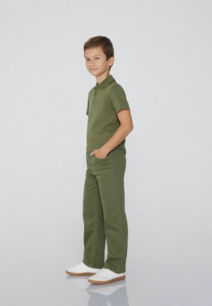 Groene polo en bijpassende groene broek, beide in een gladde stof. Witte sneakers met een bruine zool. Staande pose tegen een neutrale achtergrond.