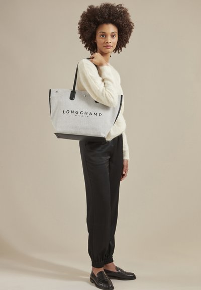 Gray felt tote bag with black leather accents, featuring the "LONGCHAMP PARIS" logo in front. Model wearing a cream sweater and black pants.