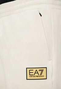 Beige trenerke iz mehkega materiala, opremljene z črno zadržno žepom in zlogom EA7 Emporio Armani na zlatem pravokotnem oznaku.