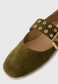 Green suede flat shoe with a square toe, featuring a wide strap with gold eyelets and a rectangular buckle. Smooth inner lining.