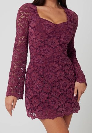 Femme portant une robe bordeaux moulante en dentelle florale à manches longues avec un décolleté carré et un ourlet et des poignets festonnés.