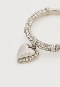 Ασημένιο βραχιόλι με υφή στη μπάντα, διακοσμημένο με διαυγή κρύσταλλα, με ένα μενταγιόν καρδιάς χαραγμένο με τη λέξη "GUESS".