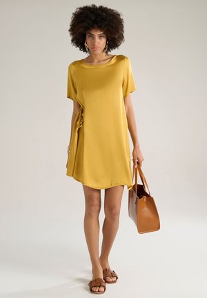 Robe en satin jaune courte avec des manches courtes, détail de lien sur le côté, et un ourlet asymétrique. Un sac à main marron et des sandales à glissière marron complètent le look.