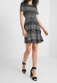 Kurzärmliges Kleid mit Schwarz-Weiß-Muster, tailliert und mit weitem Rock, kombiniert mit schwarzen High Heels.