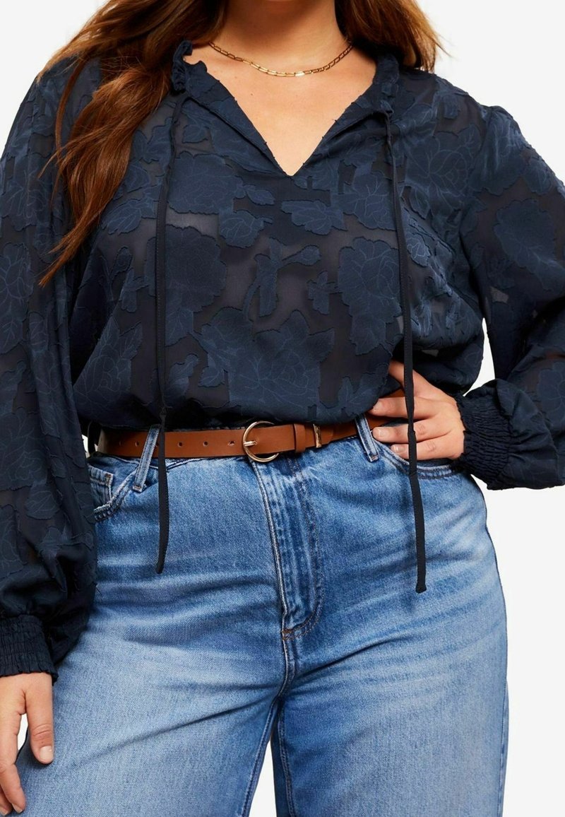 Kvinde iført gennemsigtig navyblå bluse med blomsterprint og lange ærmer, brun bælte og lyseblå jeans med høj talje, hånd på hoften, guldkæde om halsen.